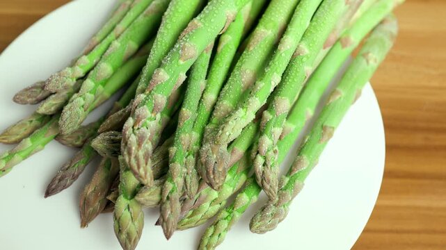Rotating fresh wet green asparagus shoots spinning slowly on a white plate on a wooden table