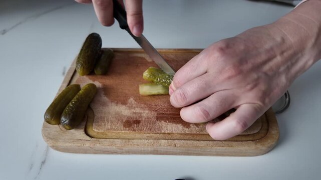 Hands slicing green pickled cucumbers with a kitchen knife on a wooden chopping board. Preparing ingredients for a salad or side dish.