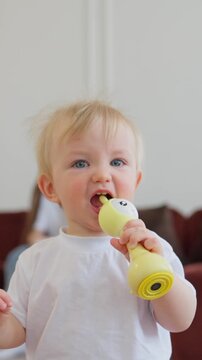 Adorable toddler with blonde hair and blue eyes chewing and playing with a yellow baby teether toy while wearing a plain white shirt in a softly lit indoor setting