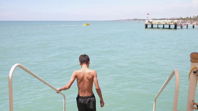 Slim young boy entering calm sea water from pier during sunny family beach holiday. Back view of teenager in dark shorts going to swim in the sea. Summer vacation concept.