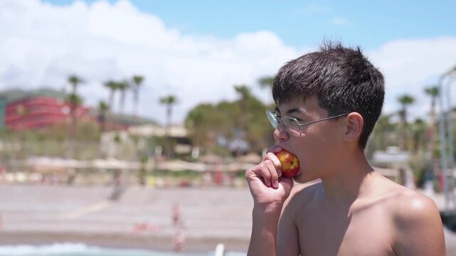 Side view of teen traveler enjoying healthy fruit snack during summer resort vacation by the sea. Teen boy with unsatisfying face eating sour peach. Unexpected facial emotions concept. Disgust refusal