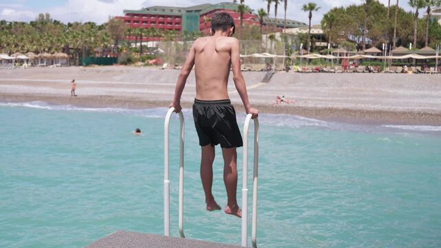 Back view of active child in dark blue shorts preparing to jump into turquoise sea water at tropical resort on sunny day. Outdoor activities at vacation at sea resort during summer holidays.