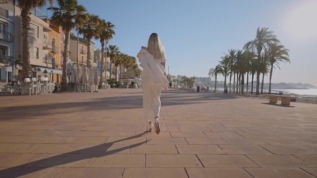 A woman walks along the waterfront in Villajoyosa. The sun is shining bright as she moves past palm trees. The street is quiet, with buildings lining the coast.