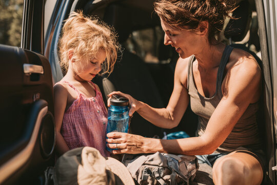 Mother giving a water bottle to a young child beside a car on a sunny summer day, representing family travel, hydration, heat safety and parenting care during road trips