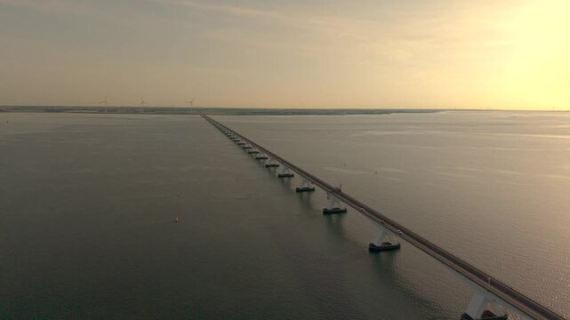 Cinematische luchtopname van de iconische Zeelandbrug tijdens golden hour. De drone vliegt vloeiend over het water terwijl auto&rsquo;s de lange brug oversteken, omringd door de rust en openheid van het Zee