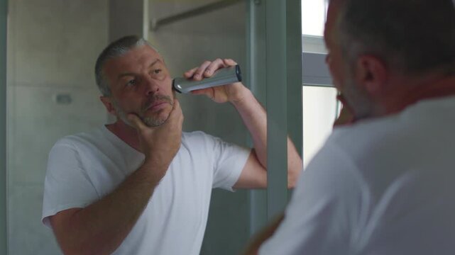 Man trimming beard in bathroom looking at mirror during morning routine