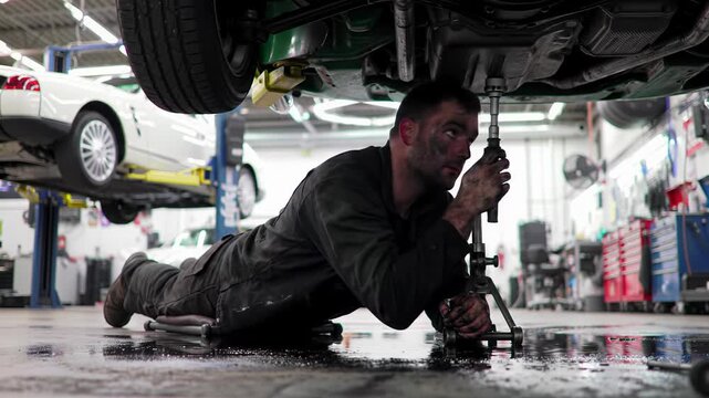 A male caucasian car mechanic in grease-stained dark work clothes lies on a creeper, using tools on a vintage emerald volkswagen karmann ghia type 14 underside in a repair shop, industrial lighting,