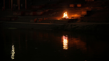 Fototapeta premium Distant sacred fire on riverside steps in dark evening atmosphere, traditional cultural ceremony, spiritual remembrance, peaceful ritual, warm flame reflection and solemn heritage