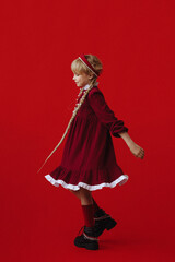 Fototapeta premium Little Caucasian girl with blonde braided hair, wearing red dress and kokoshnik, turning sideways, looking away, isolated on red background with copy space. Concept childhood, tradition, dance.