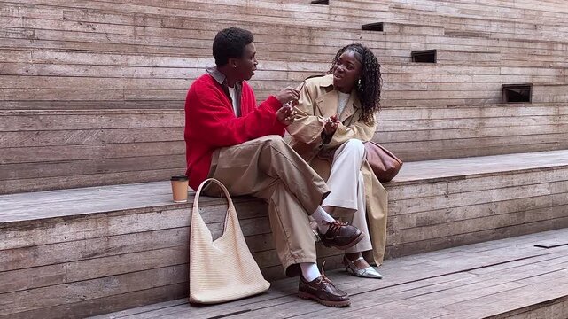 Carefree funny african american best friends man and woman sitting outdoors at public area, relaxed people black male and female on staircase talking discussing fashion accessories and joking together