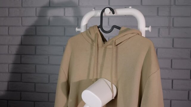 Handheld electric lint remover shaving a hoodie. Person using a modern white electric lint remover to shave pills and fuzz off a beige hooded sweatshirt hanging on a rack. Fabric care and maintenance