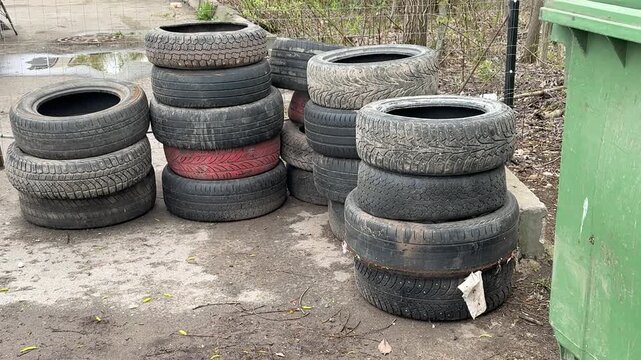 Stacked worn tires in illegal dump on street curb beside rusted fence and gravel, muddy pavement speckled with moss and red paint