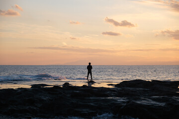Obraz premium Silhouette of a lone fisherman standing on the rocky shore of Enoshima during a calm sunset, Japan. Serene seascape near Kamakura and Tokyo. Concepts of freedom, solitude and mindfulness.
