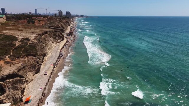 Aerial view of Sidna Ali Beach with narrow sandy shore, turquoise waves, sandstone cliffs and distant city skyline in Israel
