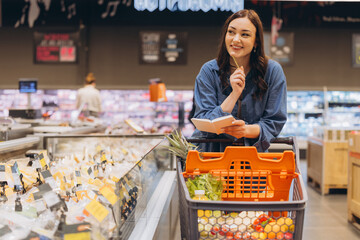 Woman holding a grocery list and pen, smiling while looking away, pushing a cart filled with fresh produce © anatoliycherkas