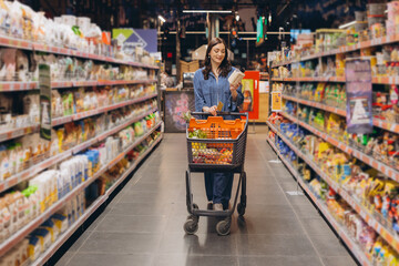 Woman shopping for groceries in supermarket aisle, pushing a full cart and checking product labels © anatoliycherkas