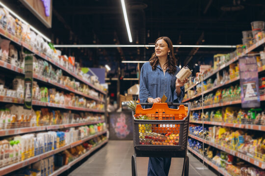 Young woman shopping for groceries in a supermarket aisle, choosing food products and pushing a full shopping cart