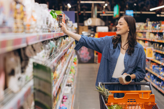 Woman shopping alone in a grocery aisle, selecting products from a shelf and pushing a cart