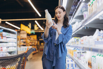 Young woman shopping for groceries, reading label on dairy bottle at supermarket, weighing options...