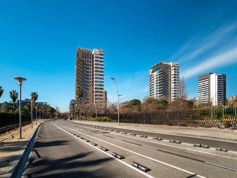 Urban landscape featuring modern skyscrapers along a clear road under a blue sky. Palm trees line the street, creating a vibrant city atmosphere.