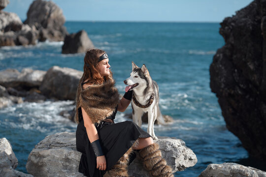 A woman poses beside a Siberian Husky dog near the sea. Rocky coastline and natural scenery create calm outdoor companionship atmosphere.
