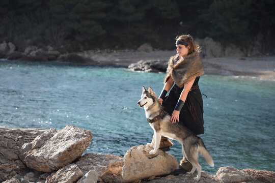 A woman stands with a Siberian Husky dog beside turquoise water. Rocky beach and scenic mountains create peaceful travel lifestyle atmosphere.