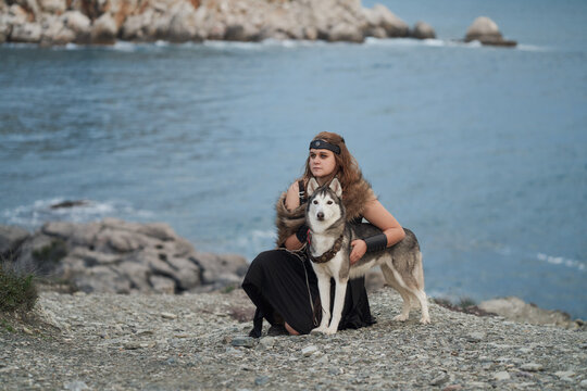 A woman and Siberian Husky dog rest together on seaside rocks. Coastal scenery with blue water and distant mountains creates cinematic outdoor adventure mood.