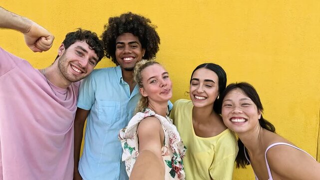 Happy group of multiethnic friends taking a selfie video against a vibrant yellow wall. Young people smiling and having fun together outdoors