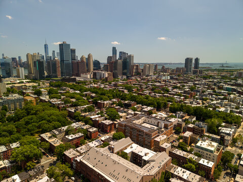 Jersey city skyline rises above a densely populated urban residential area filled with red brick buildings and lush green treetops, extending towards distant waterways on a bright summer day