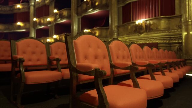 Front row red velvet seats framed by golden proscenium and warm lamp glow, ticket collector counting stubs, closeup tufted armrests and quiet anticipation