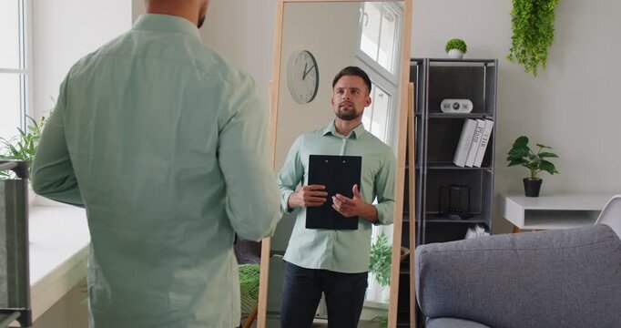 Man holds a clipboard, preparing for rehearsal and presentation, checking reflection in a mirror. Man rehearses presentation in front of mirror. Employee prepare for job conference at home