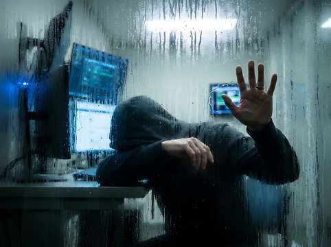 Hacker wearing a hood obscures face, resting head on hand in front of multiple computer monitors in a dark room, seen through a wet window