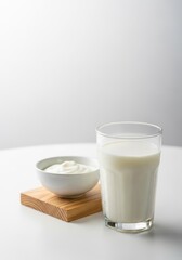 A fresh glass of cold milk stands on a white table next to a wooden block and a small bowl of creamy yogurt in a clean, minimal setting, white, refreshment, dairy