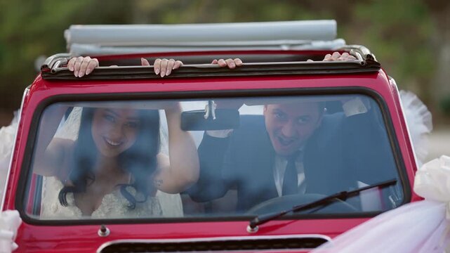 Smiling bride and groom make funny faces sitting in vintage retro convertible car during wedding celebration