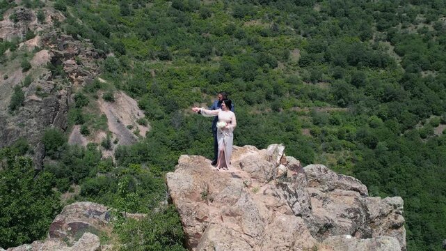 Romantic wedding couple stands on mountain peak rugged cliff holding hands and hugging surrounded by lush greenery and a mountainous landscape, enjoying a serene and romantic moment together