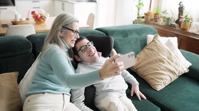 Loving mother and her adult son with a physical condition enjoying a tender moment together, taking a smiling selfie on the living room sofa