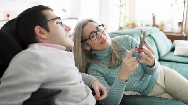Mature mother and her son with a physical condition relaxing together on a sofa at home. The woman is showing something on her smartphone