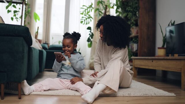 Cheerful mom spending quality time with her toddler girl at home. Little daughter holding a mobile phone and playing an educational game