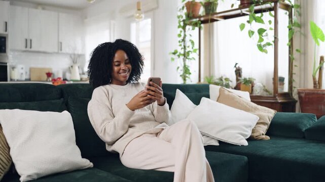 Cheerful young woman relaxing on a green sofa in a cozy living room, smiling while scrolling and typing messages on her smartphone