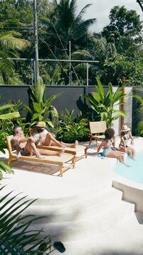 Four diverse friends enjoying a sunny day at a luxury resort. Two men are sunbathing on loungers while two women sit by the pool. Vertical