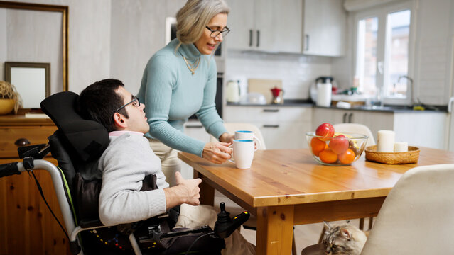 Caregiver bringing coffee to a man with a physical disability sitting in an electric wheelchair at a dining table at home
