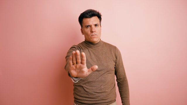 Serious man making a stop gesture, showing refusal and denial. Rejection and caution concepts against a pink background