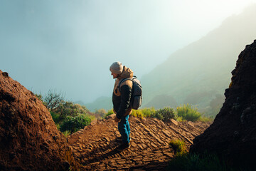 A male hiker with a backpack stands on a stone mountain path, looking out over a misty, sunlit alpine landscape. © Maria Sap