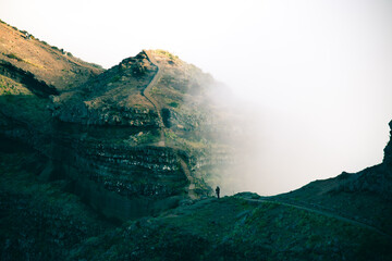 A distant hiker stands on a stone mountain path looking at a steep trail ascending a layered ridge enveloped in heavy mist. © Maria Sap