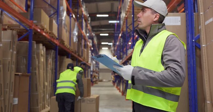 Warehouse worker checks inventory and boxes. Staff in safety vest uses a clipboard checklist to verify stock on shelves, keeping cartons counted. Reliable warehouse logistics concept today.
