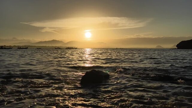 Golden sunrise over the ocean with natural wave sound. Fishing boats rest on the horizon under a glowing sky as waves roll toward the Papuma beach, Indonesia. Serene evening atmosphere with audio.