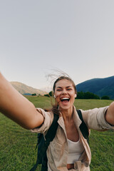 Fototapeta premium Vibrant woman outdoors, smiling with arms open, backpack ready, sunlit field and mountains behind, carefree lifestyle, candid moment, natural colors