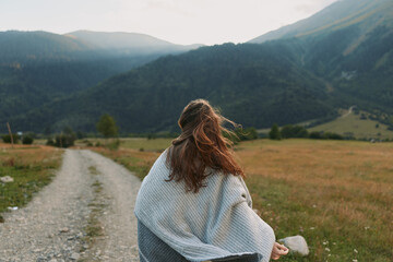Fototapeta premium Woman from behind wearing a light shawl walks along a gravel path in a mountain valley, capturing a calm countryside lifestyle moment outdoors with wind in hair