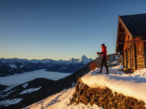 Mountaineers enjoy the sunrise in front of an alpine hut in winter, the Watzmann behind, fog in the valley, Berchtesgaden Alps, Scheibenkaser, Marktschellenberg, Berchtesgadener Land, Upper Bavaria, Bavaria, Germany