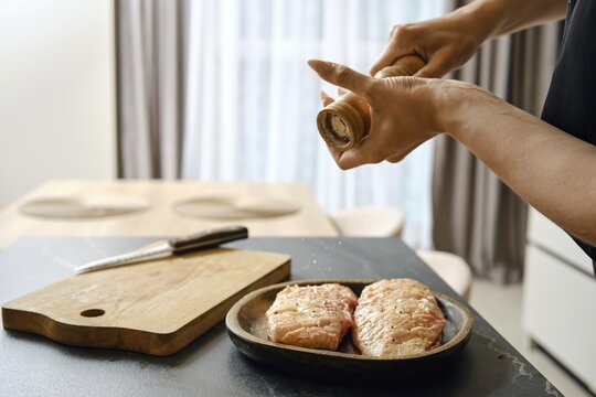 A person seasons fresh duck fillets with sea salt in a stylish kitchen. The wooden cutting board and knife are nearby, while natural light brightens the space from the window